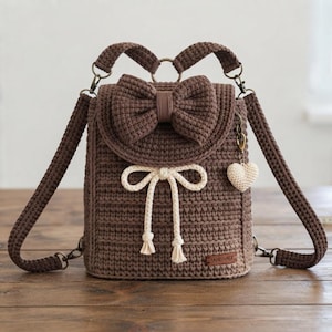 May include: A brown crocheted backpack featuring a large bow on the front flap, a cream-colored drawstring closure with tassels, and a small cream heart-shaped charm attached to the side. The bag includes adjustable straps and metal hardware.