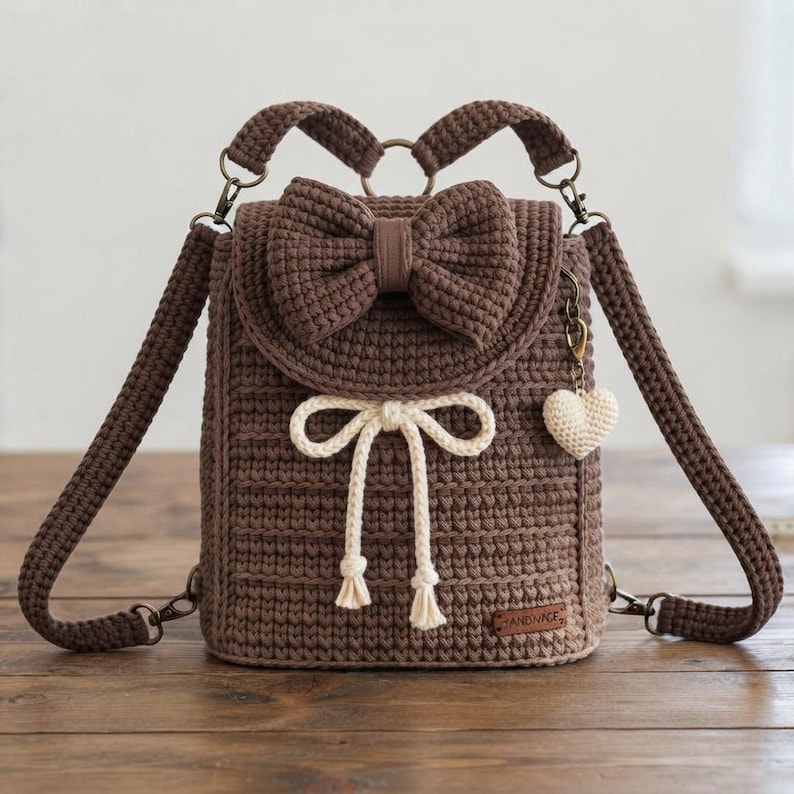 Peut inclure: Un sac &agrave; dos en crochet marron avec un grand n&oelig;ud sur le rabat avant, une fermeture &agrave; cordon cr&egrave;me avec des pompons et une petite breloque en forme de c&oelig;ur cr&egrave;me sur le c&ocirc;t&eacute;. Le sac comprend des bretelles r&eacute;glables et des pi&egrave;ces m&eacute;talliques.