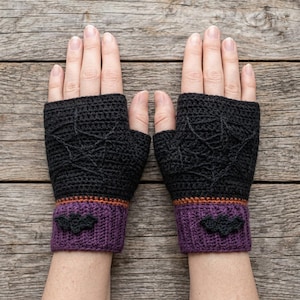 Puede incluir: Un par de guantes sin dedos tejidos a ganchillo con hilo negro que presentan un patrón de telaraña en el dorso de la mano. Los puños son morados con una fina raya naranja y una pequeña aplicación de murciélago negro en la muñeca.