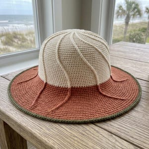 Peut inclure: Un chapeau de soleil en crochet composé d'une calotte crème avec des nervures verticales en relief qui se prolonge par un large bord terracotta. Le bord est fini par une fine bordure vert olive foncé, créant un contraste de couleurs net.