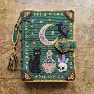 May include: A dark green crochet book cover featuring a crescent moon, a black cat, a potion bottle, a skull with crystals, and a raven. The cover is decorated with embroidered stars, runic text, and brown corner accents with a metal buckle closure.