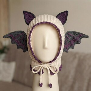 May include: A cream-colored crochet balaclava featuring two pointed ears and two large, dark gray bat wings extending from the sides. The hood is trimmed with a purple scalloped edge and includes two long, matching cream-colored ties at the chin.