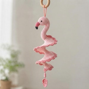 Pode incluir: Um móbile suspenso de flamingo em crochê rosa claro com um design de corpo em espiral e bordas festonadas. O flamingo tem um pequeno olho preto e um bico escuro. Está suspenso por um anel de madeira clara com uma alça de fio rosa e finalizado com um pequeno pendente de crochê em forma de folha.