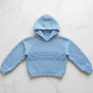 May include: A light blue long-sleeve hooded sweater crafted with a crochet technique. The garment features a textured stitch pattern across the chest and sleeves, a hood, and ribbed cuffs at the wrists and hem, providing a consistent solid color.