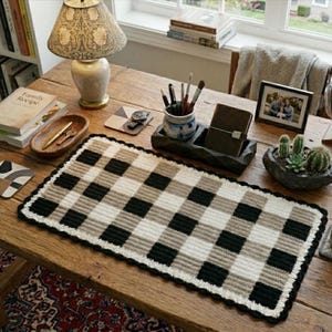 May include: A rectangular crochet desk mat featuring a repeating buffalo plaid pattern in black, white, and tan. The mat has a textured, ribbed surface and a scalloped black border, positioned centrally on a wooden desk surface with various office items.
