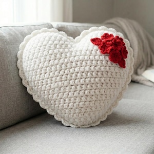 May include: A cream-colored, heart-shaped crochet pillow with a scalloped edge and a cluster of red crochet flowers on the upper right side. The pillow is placed on a gray sofa, showcasing its handmade texture and design.