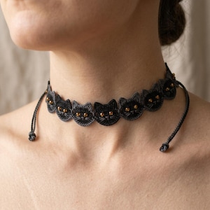 Könnte beinhalten: Eine schwarze Häkel-Halskette mit einem sich wiederholenden Muster aus Katzengesichtern. Jedes Katzengesicht ist aus dunklem Garn gefertigt und hat kleine, glänzende goldfarbene Perlen als Augen. Die Kette ist mit dünnen schwarzen Bändern verstellbar.