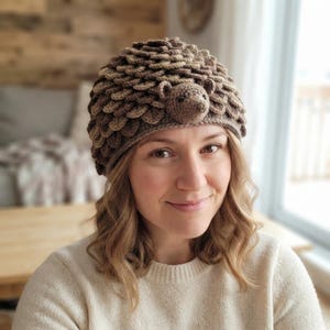 May include: A brown crochet beanie featuring a textured, layered scale pattern that mimics the appearance of a hedgehog. The hat includes a small, three-dimensional crocheted hedgehog face with ears and a snout centered on the front brim.