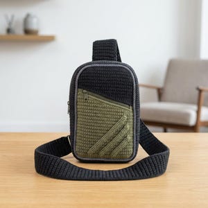 May include: A black and olive green crochet sling bag featuring a single wide shoulder strap, a zippered front pocket, and three decorative diagonal crochet lines on the lower front section. The bag has a structured rectangular shape.
