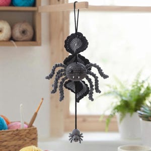 May include: A handmade crochet hanging decoration featuring a large dark gray spider with eight legs, two white fangs, and black eyes, suspended above a smaller matching gray spider, both attached to a black string with a decorative black web element.