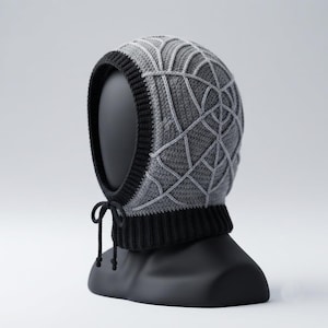 Pode incluir: Uma balaclava de crochê cinza com um padrão de teia de aranha preta no topo. A peça inclui uma borda canelada preta espessa ao redor da abertura do rosto e uma base de pescoço canelada preta combinando com dois cordões pretos finos.