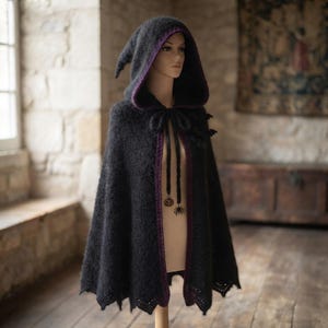 May include: A black hooded cape with a fuzzy texture and a purple inner lining. The cape features a pointed hood, a front tie closure with dangling charms shaped like a pumpkin and a spider, and a scalloped hemline with a delicate lace pattern.