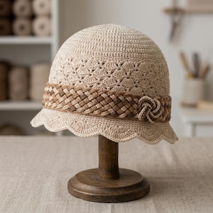 May include: A beige crochet bucket hat featuring a textured shell stitch pattern on the crown and brim. A wide, brown braided band encircles the base of the crown, accented with a small, matching braided knot detail on the side of the hat.