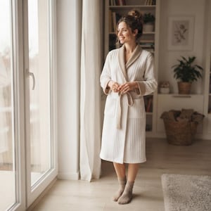 Op de afbeelding: Een knielange, geribbelde witte badjas met een contrasterende beige sjaalkraag, bijpassende beige boorden en een ceintuur. De badjas is voorzien van twee opgestikte zakken aan de voorzijde en lange mouwen voor een comfortabele pasvorm.