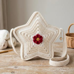 May include: A star-shaped crochet bag in cream yarn with a central dark red flower appliqué featuring a yellow centre. The bag includes a matching crochet shoulder strap and a small wooden bead accent attached to the side near the strap connection.