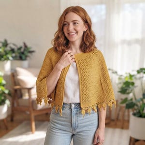 May include: A mustard yellow crochet capelet featuring a textured stitch pattern and a decorative fringe along the bottom hem. The garment is worn over a simple white top and light blue denim jeans, showcasing a classic open-front design.