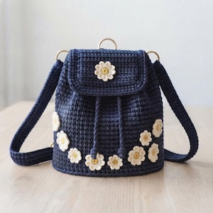 Puede incluir: Una mochila de ganchillo en color azul marino oscuro con cierre de cordón y solapa superior. El bolso está decorado con varios apliques de flores de margarita tejidas en blanco y amarillo. Incluye dos correas y un asa superior pequeña.