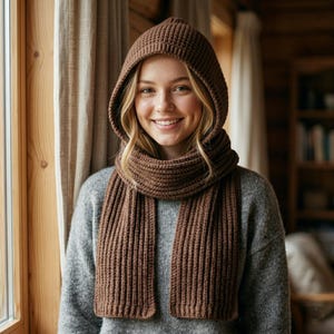 Könnte beinhalten: Ein brauner Kapuzenschal mit gerippter Strickstruktur. Das Kleidungsstück besteht aus einer passgenauen Kapuze, die in einen langen, integrierten Schal übergeht, der in zwei vertikalen Bahnen herabhängt, gefertigt aus braunem Garn mit gleichmäßigem Maschenbild.