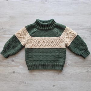 May include: A long-sleeve crochet sweater featuring a dark forest green body and sleeves with a contrasting cream-colored horizontal band across the chest and shoulders, decorated with a raised diamond-shaped cable stitch pattern and a ribbed collar.