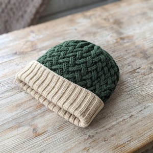 Può includere: Un berretto in maglia caratterizzato da una corona verde bosco scuro con un motivo a intreccio testurizzato e un ampio risvolto piegato in un colore beige chiaro a contrasto. Il berretto presenta una lavorazione a coste sul risvolto e una trama fitta.