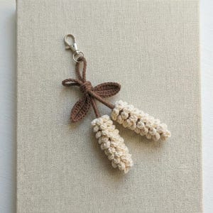May include: A handmade crochet keychain with two off-white, textured, cylindrical elements. The keychain has a brown bow and leaves, and a silver-coloured clasp. The item is set against a neutral background.