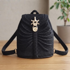 Puede incluir: Una mochila de ganchillo negra con un diseño inspirado en una caja torácica en la solapa frontal. El cierre está asegurado con un adorno de color crema en forma de hueso y un botón circular a juego, aportando un detalle estructural único.