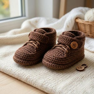 May include: A pair of brown crochet baby booties. The booties have laces, a button closure, and a velcro strap. The shoes are made of a textured yarn and are sitting on a cream-colored surface.