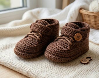 May include: A pair of brown crochet baby booties. The booties have laces, a button closure, and a velcro strap. The shoes are made of a textured yarn and are sitting on a cream-colored surface.