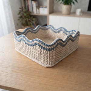 May include: A square, hand-crocheted basket made from thick, cream-colored yarn. The basket features a distinct wavy, scalloped top edge accented with a contrasting dusty blue yarn trim, creating a textured, structured container for storage.
