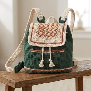Puede incluir: Una mochila de ganchillo en verde oscuro, crema y terracota. La bolsa cuenta con una solapa con patrón trenzado, un bolsillo frontal, dos correas de hombro color crema, un asa superior y un cierre de cordón con dos borlas crema colgando.