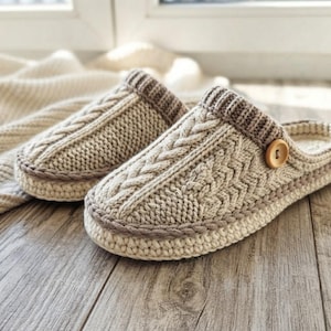 Peut inclure: Une paire de chaussons en tricot beige avec un motif torsadé sur le dessus, un revers côtelé et un bouton en bois marron clair sur le côté. Les chaussons présentent une bordure crochetée épaisse autour de la base et une texture tricotée.