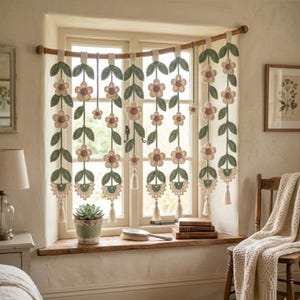Può includere: Una mantovana decorativa per finestra composta da diversi fili all'uncinetto pendenti con fiori color crema, foglie verde salvia e piccole nappe sul fondo. I fili sono sospesi a un bastone in legno curvo montato sulla finestra.