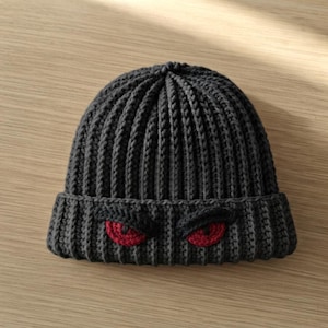 May include: A dark charcoal grey knit beanie featuring a ribbed texture and a folded cuff. The cuff displays two embroidered red eyes with black pupils, positioned side by side, creating a distinct graphic detail on the front of the hat.