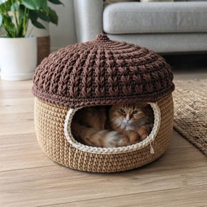 Pode incluir: Uma cama de crochê para animais de estimação em forma de bolota, com uma tampa texturizada castanho-escura e uma base bege-clara. A cama apresenta uma abertura larga e arredondada com uma borda creme, com um gato tigrado laranja no interior.