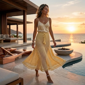 May include: A woman wearing a cream-colored silk camisole top and a yellow crochet skirt with a drawstring waist and tassel detail. The skirt has a flared hem with small pom-poms. The background features a sunset over the ocean.