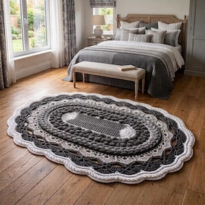 May include: An oval-shaped crochet rug featuring concentric layers of dark gray, light gray, and white yarn. The design incorporates textured bobble stitches in the center, surrounded by intricate openwork patterns and a scalloped white border.