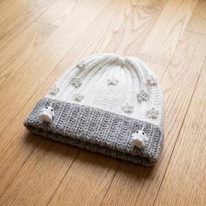 May include: A white and gray ribbed knit beanie featuring small crocheted star appliques scattered across the white crown. The gray folded brim is decorated with two small, white, ghost-shaped crocheted figures with black bead eyes.