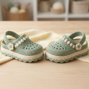 May include: A pair of sage green crochet clogs featuring a thick cream-colored sole with a scalloped edge. Each shoe has a matching cream-colored strap adorned with a small flower detail on the side and a perforated top design.