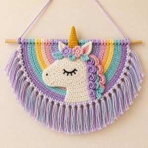 Op de afbeelding: Een gehaakte wandhanger met een crèmekleurige eenhoornkop met een gouden hoorn, bloemaccenten en een veelkleurige manen. De eenhoorn is gecentreerd op een regenboogvormige achtergrond met pastelkleurige strepen en lange, lichtpaarse franjes.