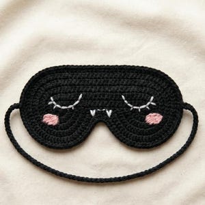 Puede incluir: Un antifaz de crochet negro con ojos cerrados bordados en blanco con pestañas, dos pequeños colmillos blancos en el centro y dos mejillas circulares de color rosa. El antifaz incluye una fina correa de crochet negra para sujetarlo.