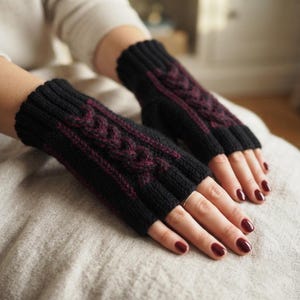 May include: A pair of fingerless gloves featuring a black knit base with a prominent dark burgundy cable knit pattern running vertically along the center. The gloves have a ribbed cuff at the wrist and a ribbed opening for the fingers.
