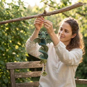 May include: A green crocheted dragon hanging ornament with a spiral body, small wings, and a tail ending in a clawed foot. The dragon features a textured yarn surface, small horns, and a string of colourful beads attached near the bottom tail section.