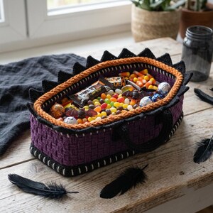 May include: A rectangular purple crocheted basket with a black serrated rim and an orange inner border. The basket is filled with assorted candies, including jelly beans, chocolate bars, and foil-wrapped treats, along with small plastic bat figures.