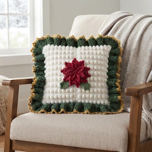 May include: A square crochet pillow featuring a cream-colored bobble stitch center with a central red poinsettia flower and green leaves. The pillow is bordered by a dark green ruffled edge trimmed with a thin gold-colored yarn accent.