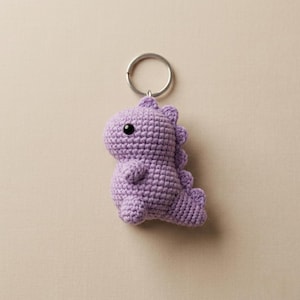 May include: A lavender crocheted dinosaur keychain with a silver metal ring. The dinosaur has a textured surface, a black eye, and a cute, friendly design. The keychain is a handmade item.