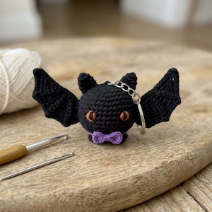 May include: A black crocheted bat keychain featuring large orange eyes and a small purple bow tie. The bat has two pointed ears and wide, textured wings. A silver metal keyring is attached to the top of the bat's head, providing a functional loop.