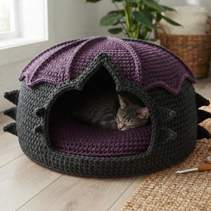 Puede incluir: Una cueva para mascotas tejida a ganchillo en gris oscuro y morado con un diseño inspirado en un dragón, con aletas laterales puntiagudas y un techo festoneado. Un gato atigrado duerme acurrucado dentro sobre un cojín morado.