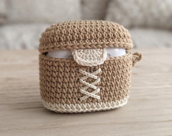 May include: A tan crochet protective case for wireless earbuds featuring a cream-colored lace-up design on the front. The case has a matching tan lid and a cream-colored base trim, with a small cream-colored tab detail centered below the lid.