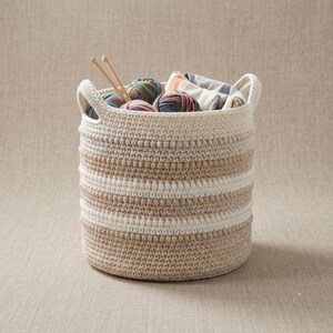 May include: A round, crocheted storage basket featuring horizontal stripes in cream, beige, and tan. The basket contains several multicolored yarn balls and two wooden knitting needles protruding from the top, creating a textured, functional container.