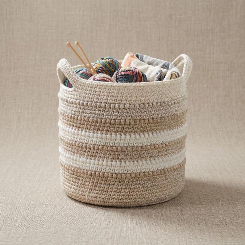 May include: A round, crocheted storage basket featuring horizontal stripes in cream, beige, and tan. The basket contains several multicolored yarn balls and two wooden knitting needles protruding from the top, creating a textured, functional container.